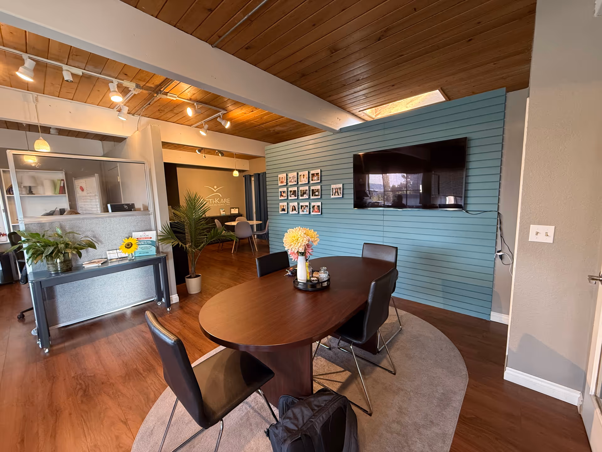Interior lounge area with oval table, chairs, TV on teal wall, plants, and reception desk.