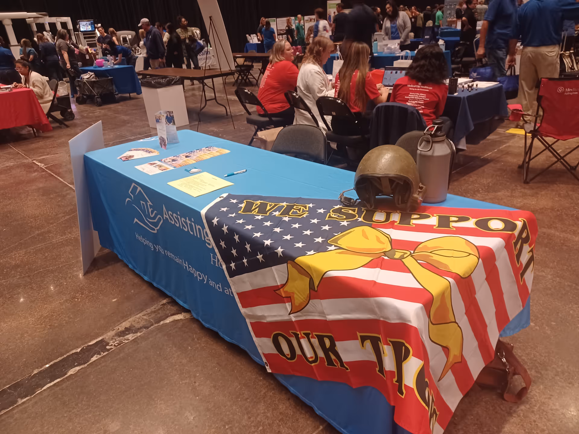 Indoor event hall with blue-covered tables; Assisting Hands Home Care banner visible.