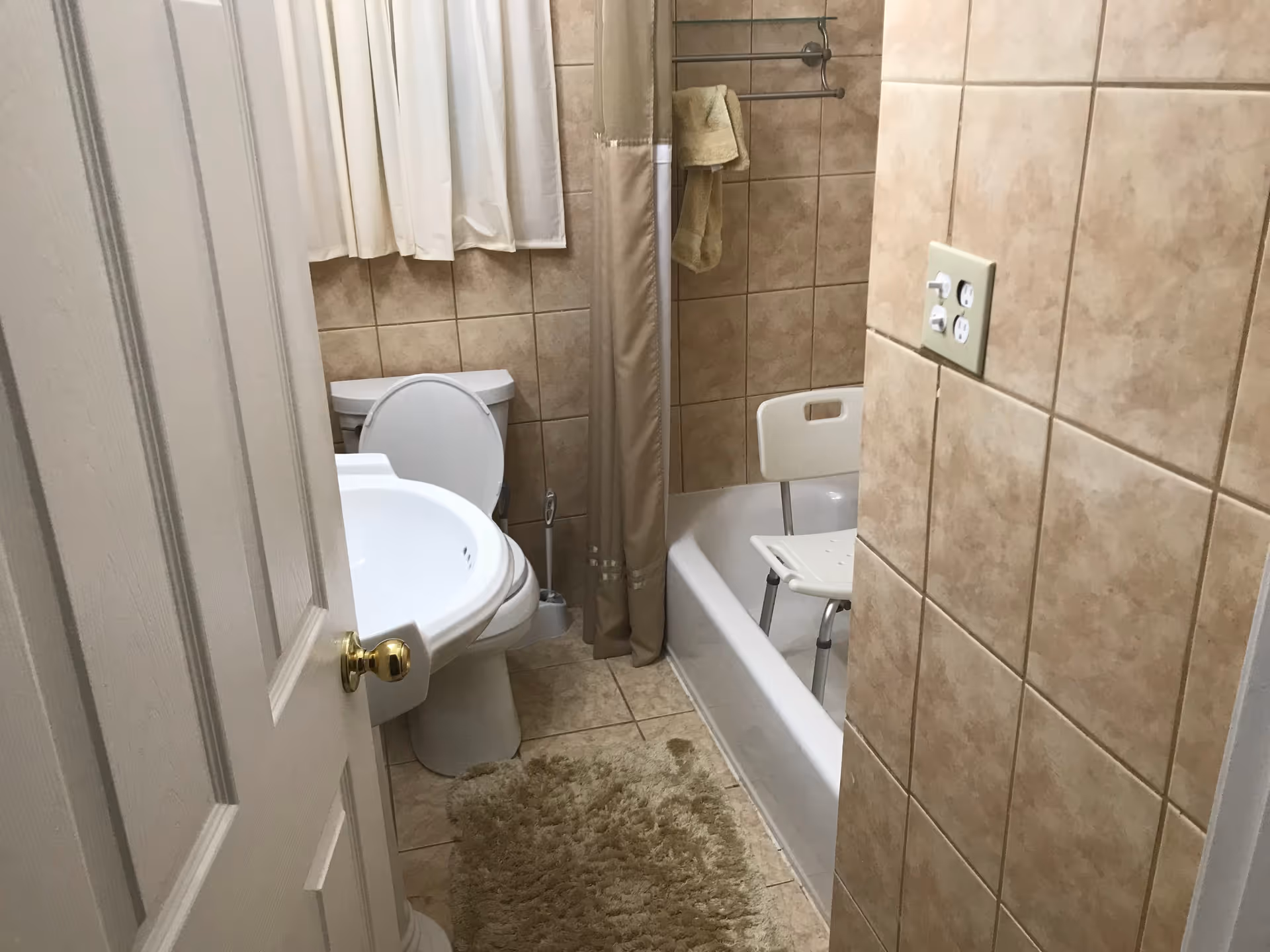 Small beige-tiled bathroom with sink, toilet, tub, shower curtain, and rug