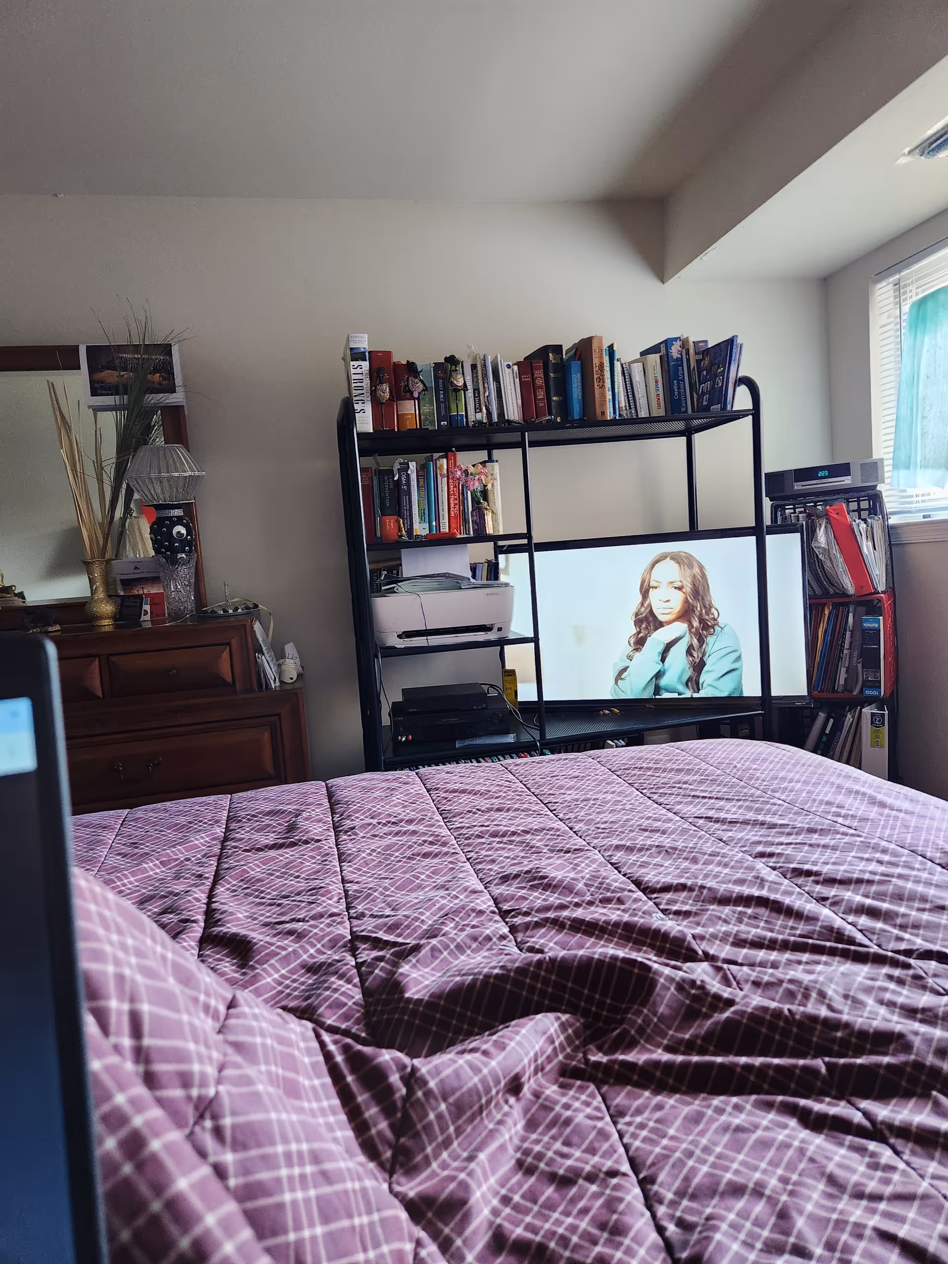 Bedroom scene with a purple quilted bedspread, a TV stand with a screen, and a window.