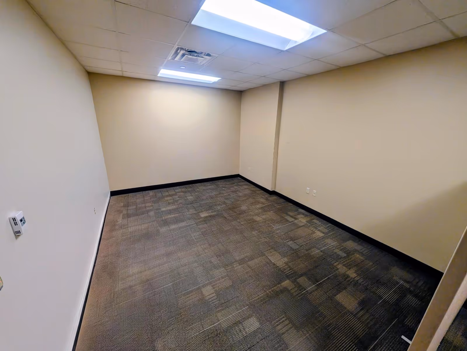 Empty beige-walled interior room with carpet, ceiling tiles, and fluorescent lights.