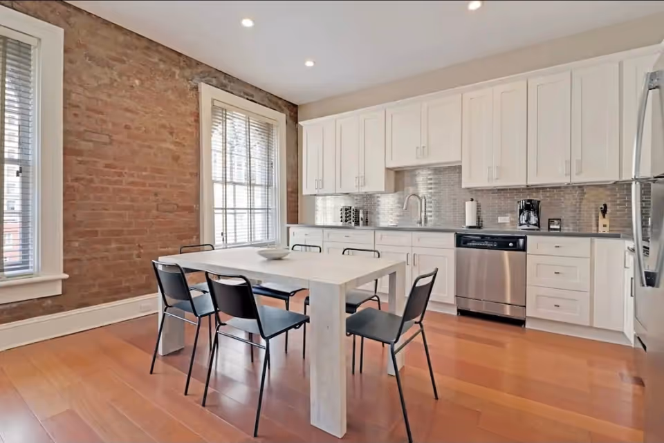Bright modern kitchen with white cabinets, stainless appliances, brick accent wall, and a dining table.