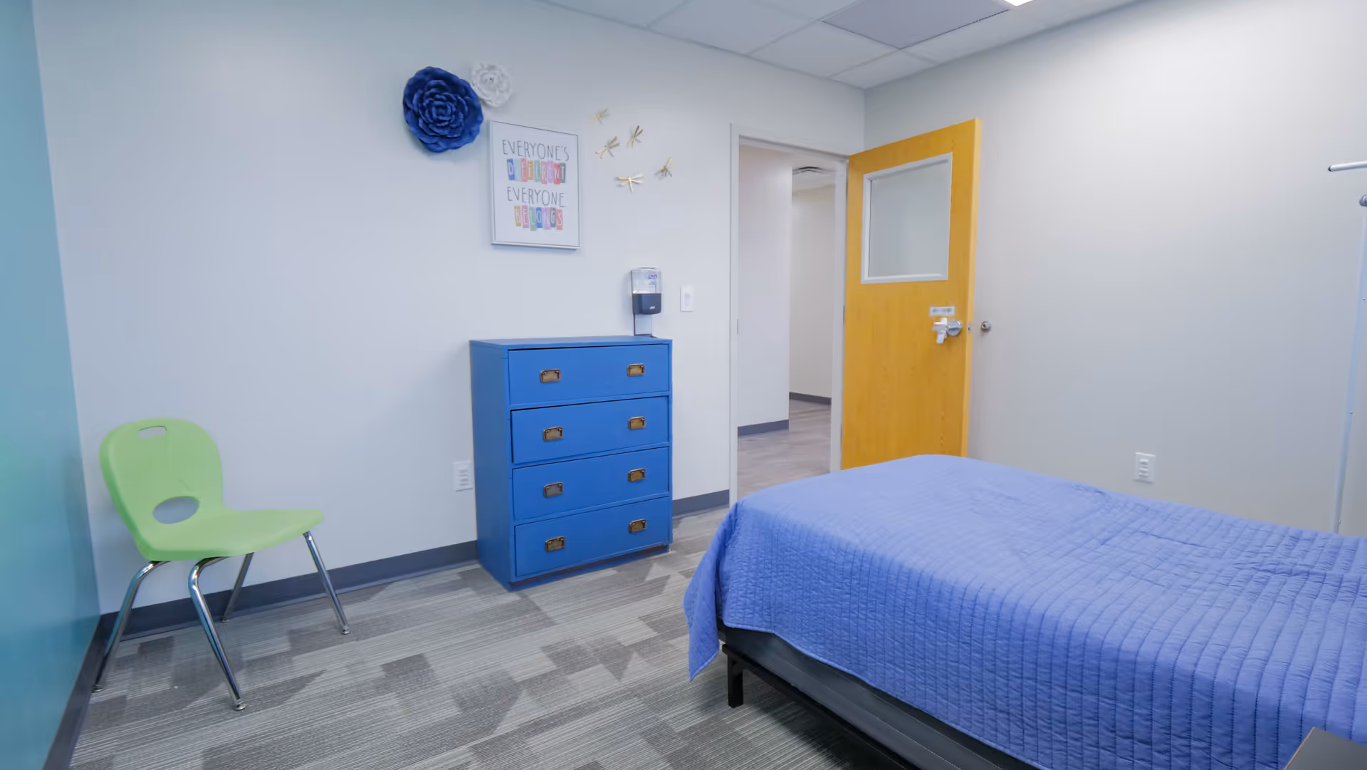 A neat bedroom in a senior living facility with a blue bedspread, blue dresser, and chair.