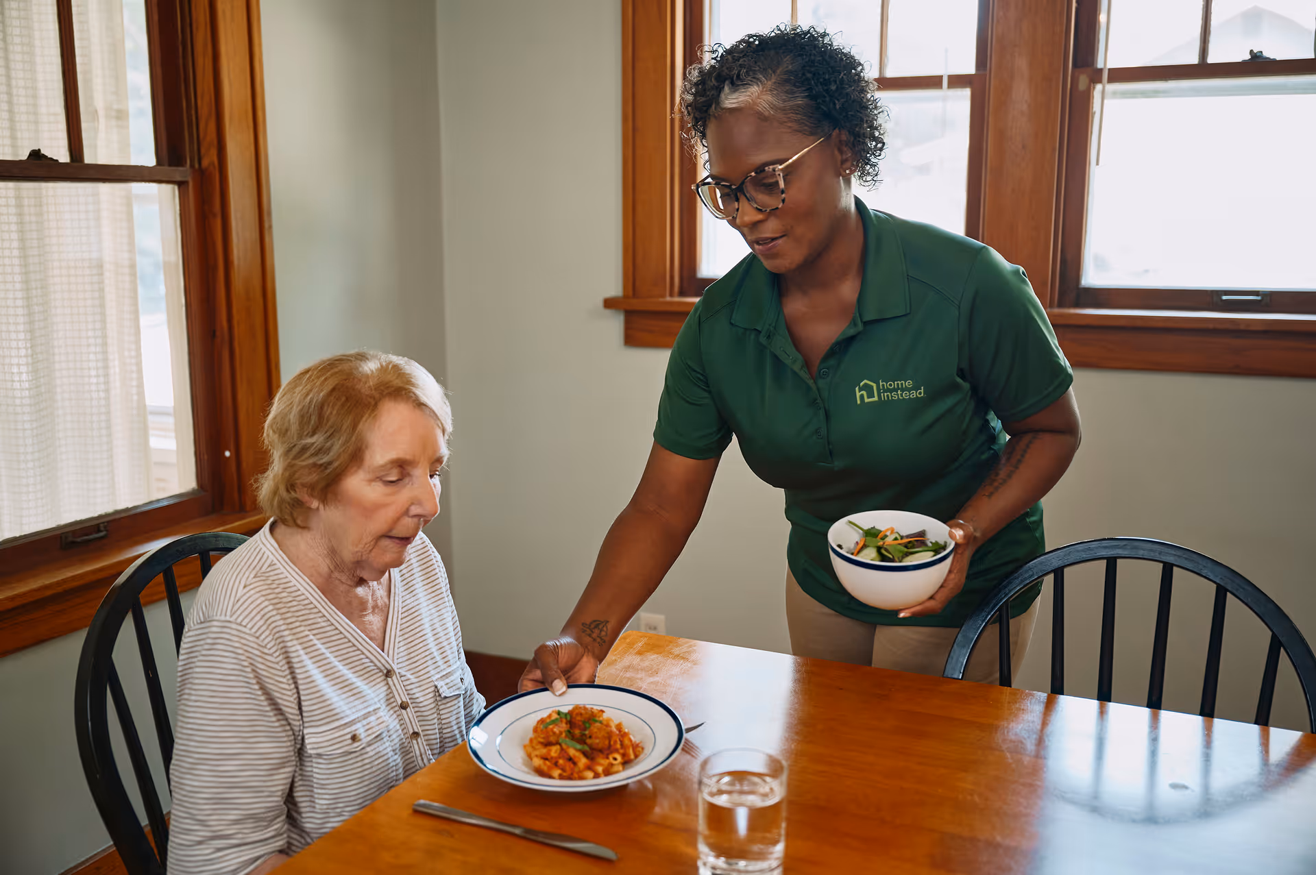 Caregiver in green Home Instead shirt serves a meal to an elderly woman at a table.