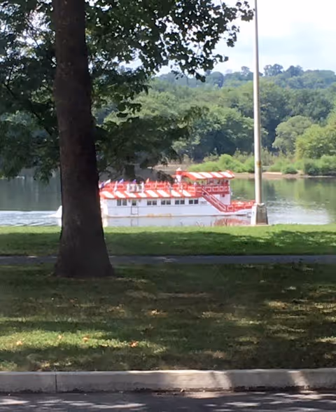 Red and white riverboat on calm water seen from a grassy park by trees