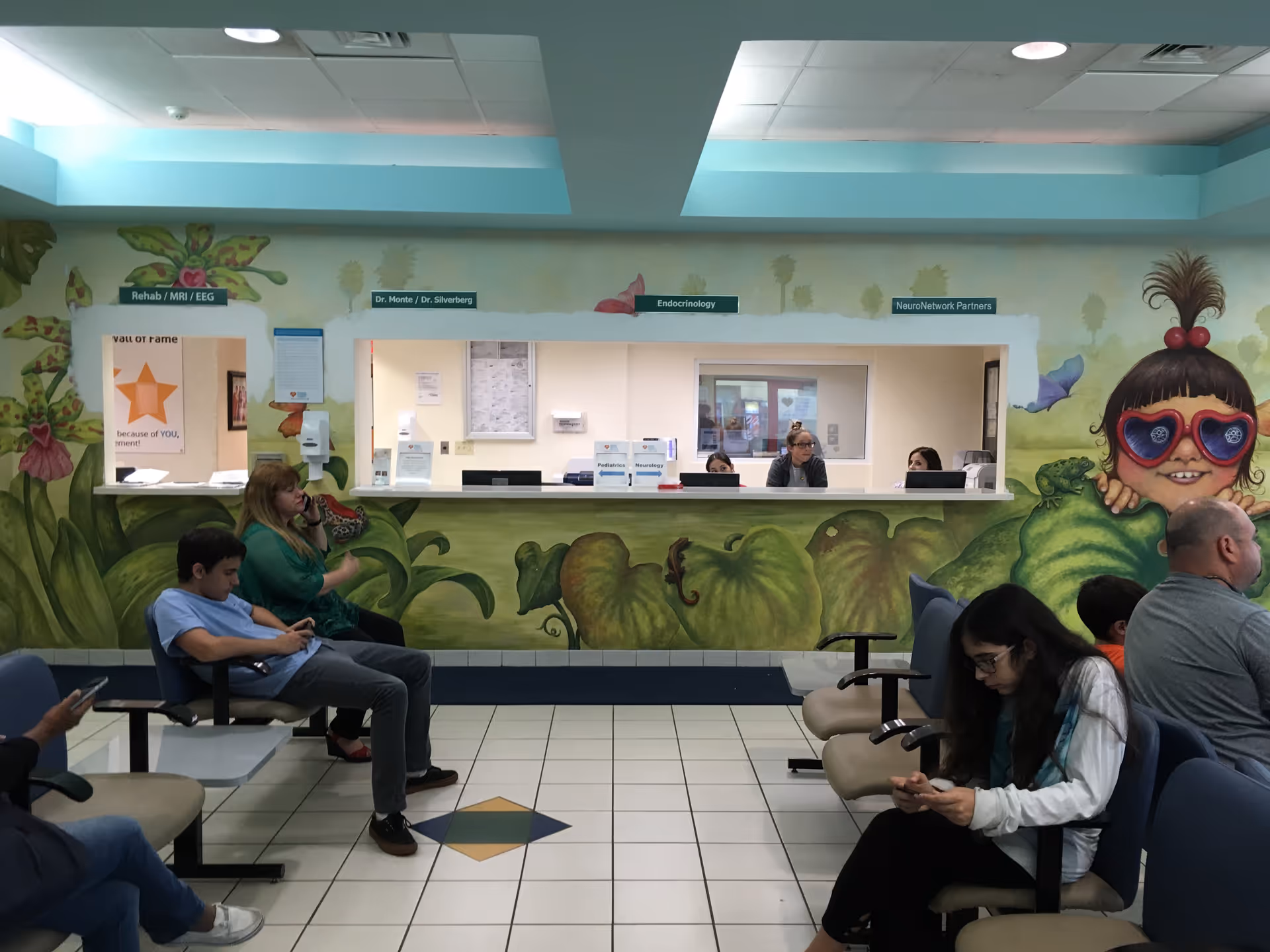 Hospital lobby with a colorful mural, reception desk, and people seated in chairs.
