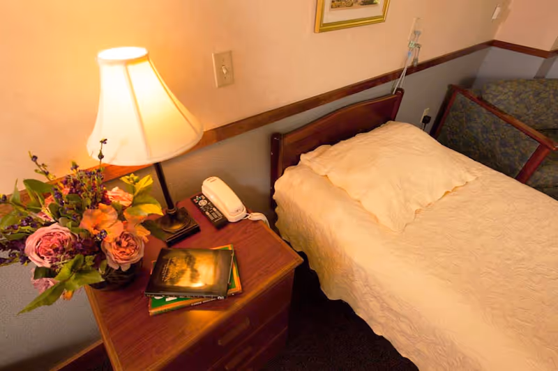 A cozy bedroom with a bed, nightstand, lamp, flowers, and books.
