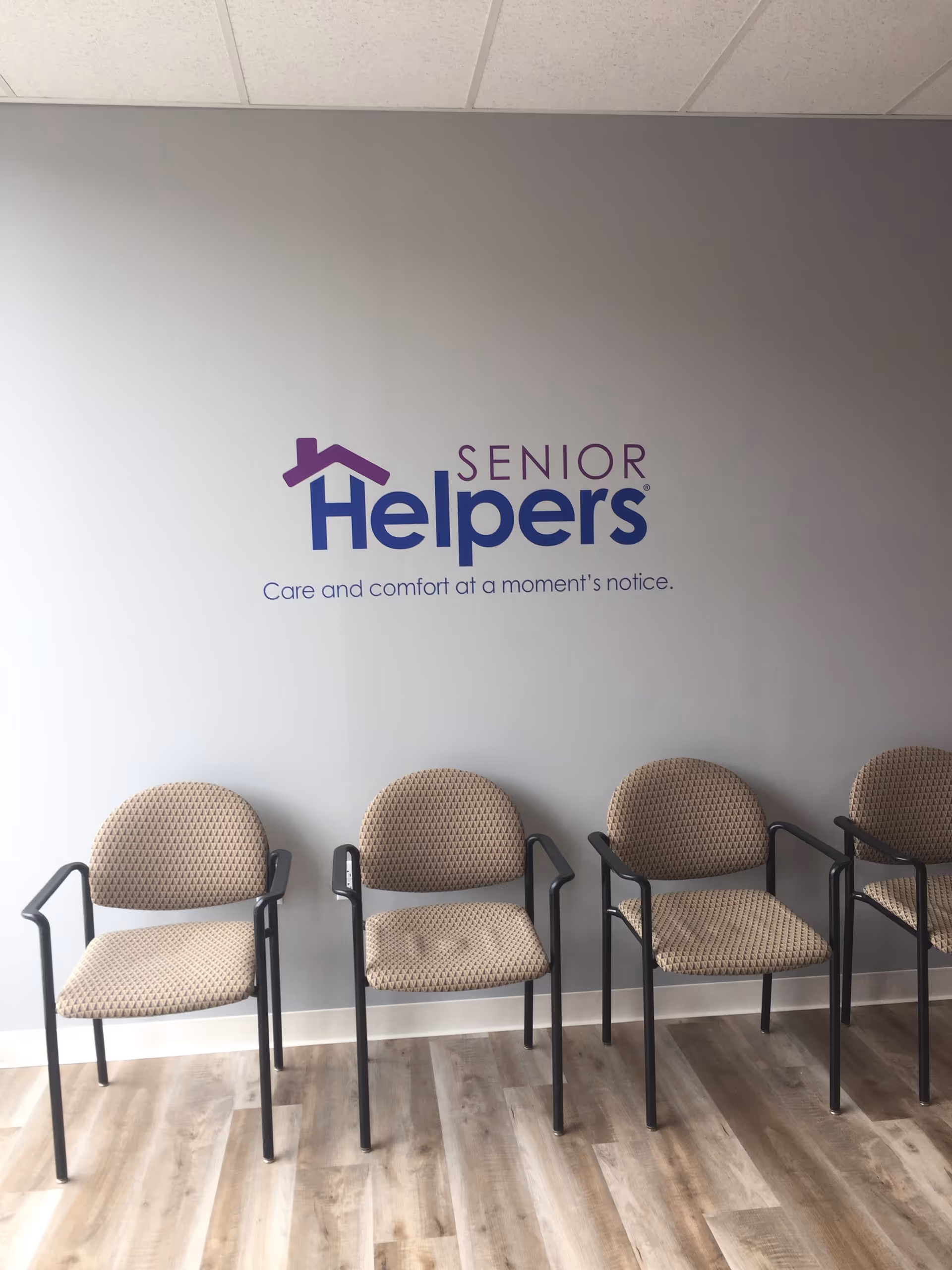 Interior waiting area with a row of beige chairs and a Senior Helpers logo on wall.