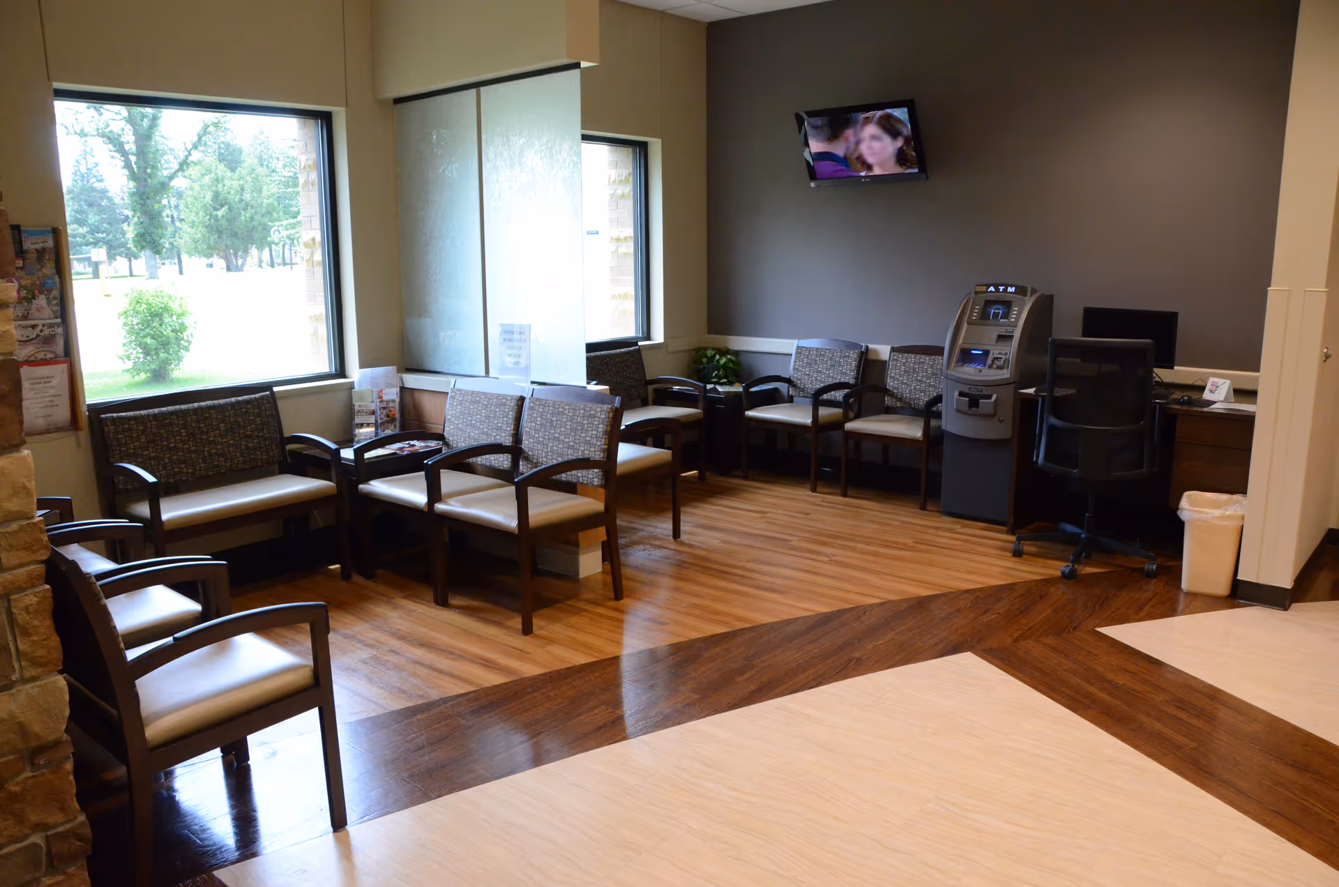 Sunlit waiting area with chairs along windows, a desk, TV, and an ATM-like kiosk.