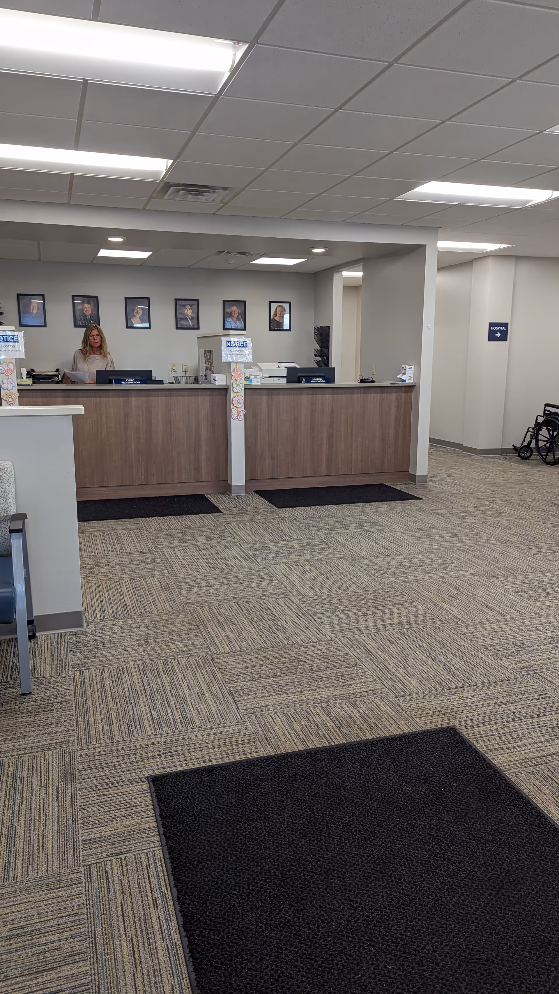 Lobby reception desk with a staff member, wall portraits, and a wheelchair nearby.