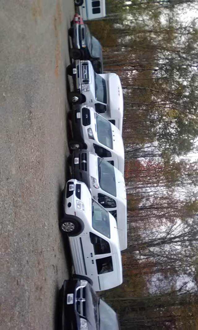Row of white vans parked in an outdoor lot with trees in the background.