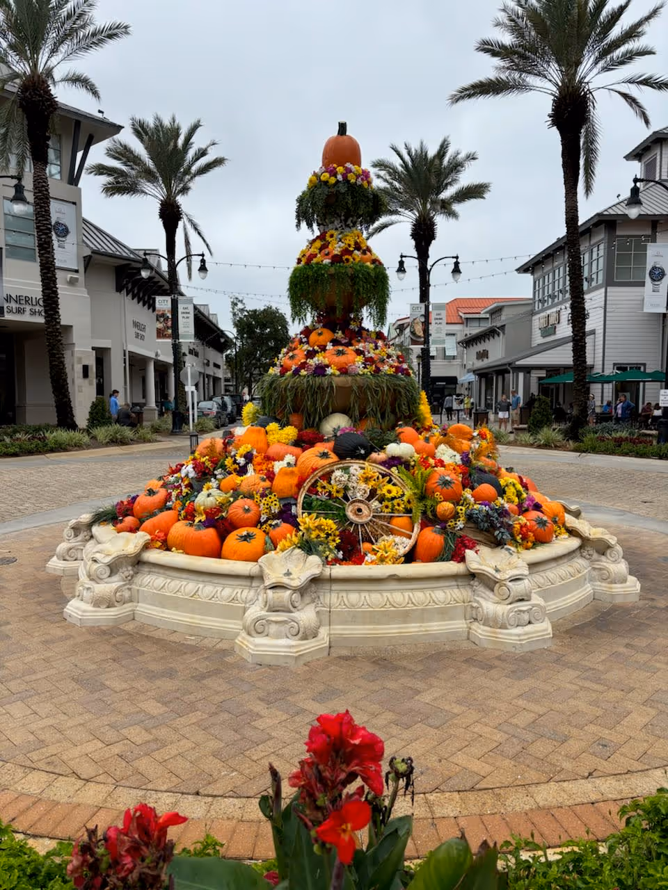 A tall pumpkin and flower tower surrounded by pumpkins in an outdoor shopping plaza.