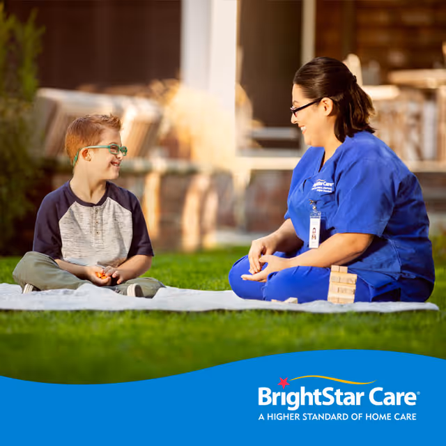 Caregiver in blue scrubs sits on a blanket outside with a smiling boy.