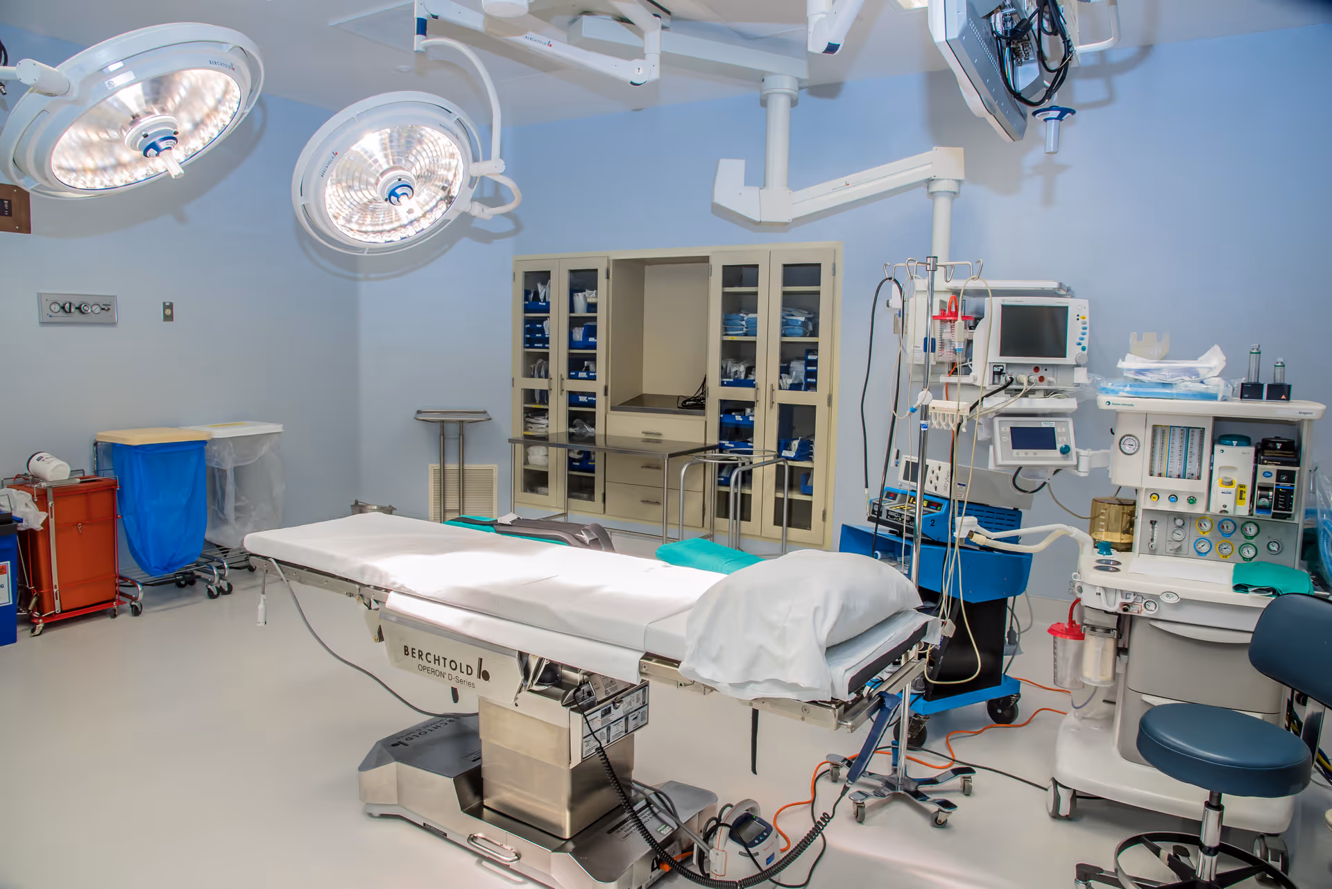 Sterile operating room with surgical table, overhead lights, and anesthesia equipment.