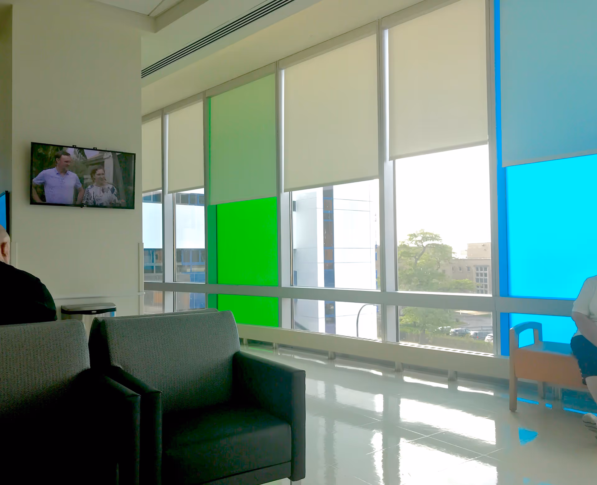 Bright modern waiting area with large windows, colored panels, grey couches, and a wall-mounted TV.
