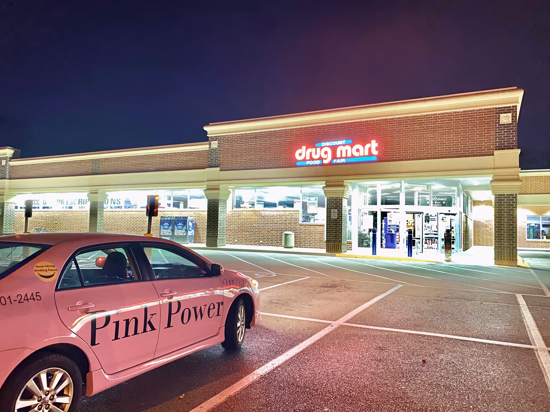 Nighttime exterior of Discount Drug Mart with a pink 'Pink Power' car parked outside.