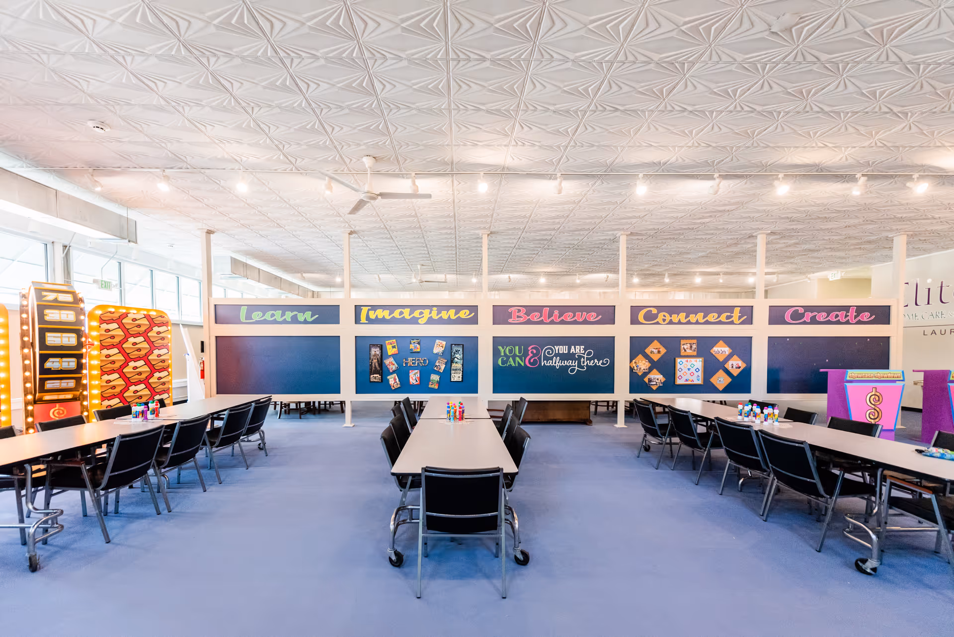 Bright multipurpose interior with long tables and chairs, colorful wall panels reading Learn, Imagine, Believe, Create.