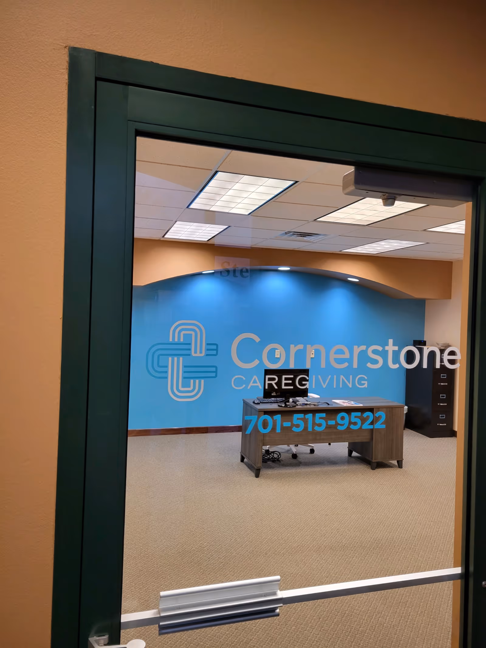 Glass door reveals Cornerstone Caregiving logo, blue wall, and desk with computer inside an office.