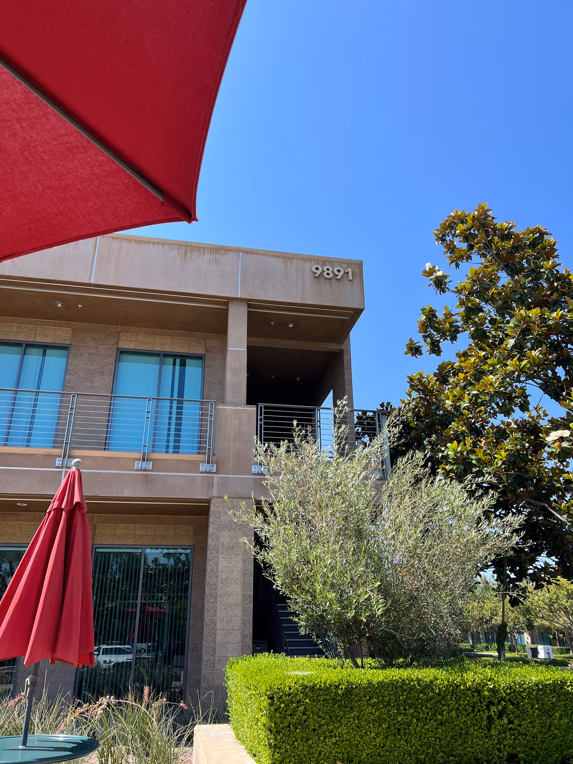 Two-story tan building with balcony, red patio umbrella, blue sky, and landscaped foreground.