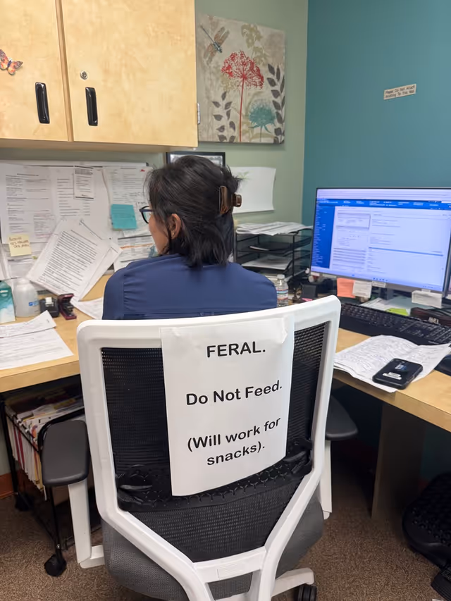 Office worker seated at desk with sign 'FERAL. Do Not Feed' on chair.
