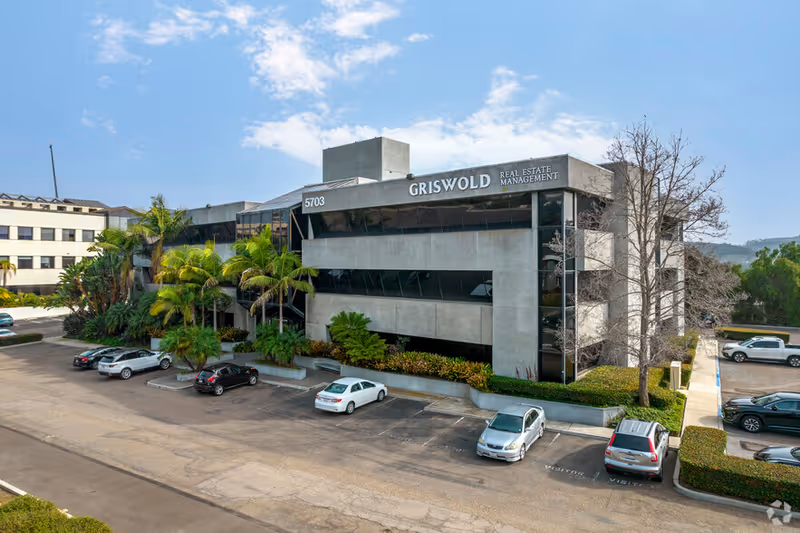 Modern office building with parking lot, palm trees, and blue sky.