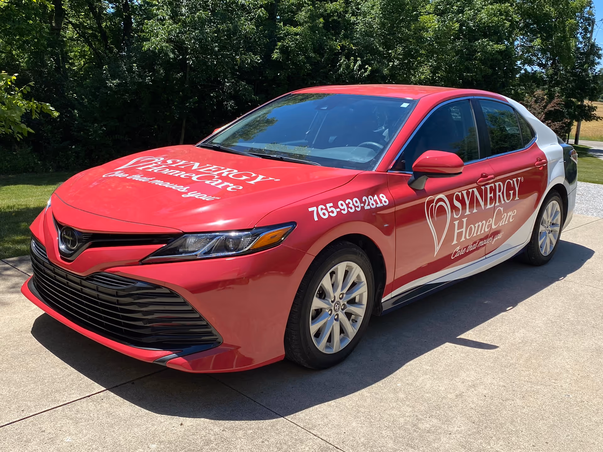 Red Synergy HomeCare car with logo parked on a driveway beside trees.