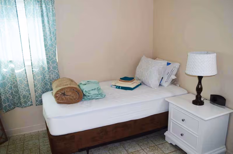 A small bedroom with a single bed, folded linens, a nightstand, lamp, and curtains.