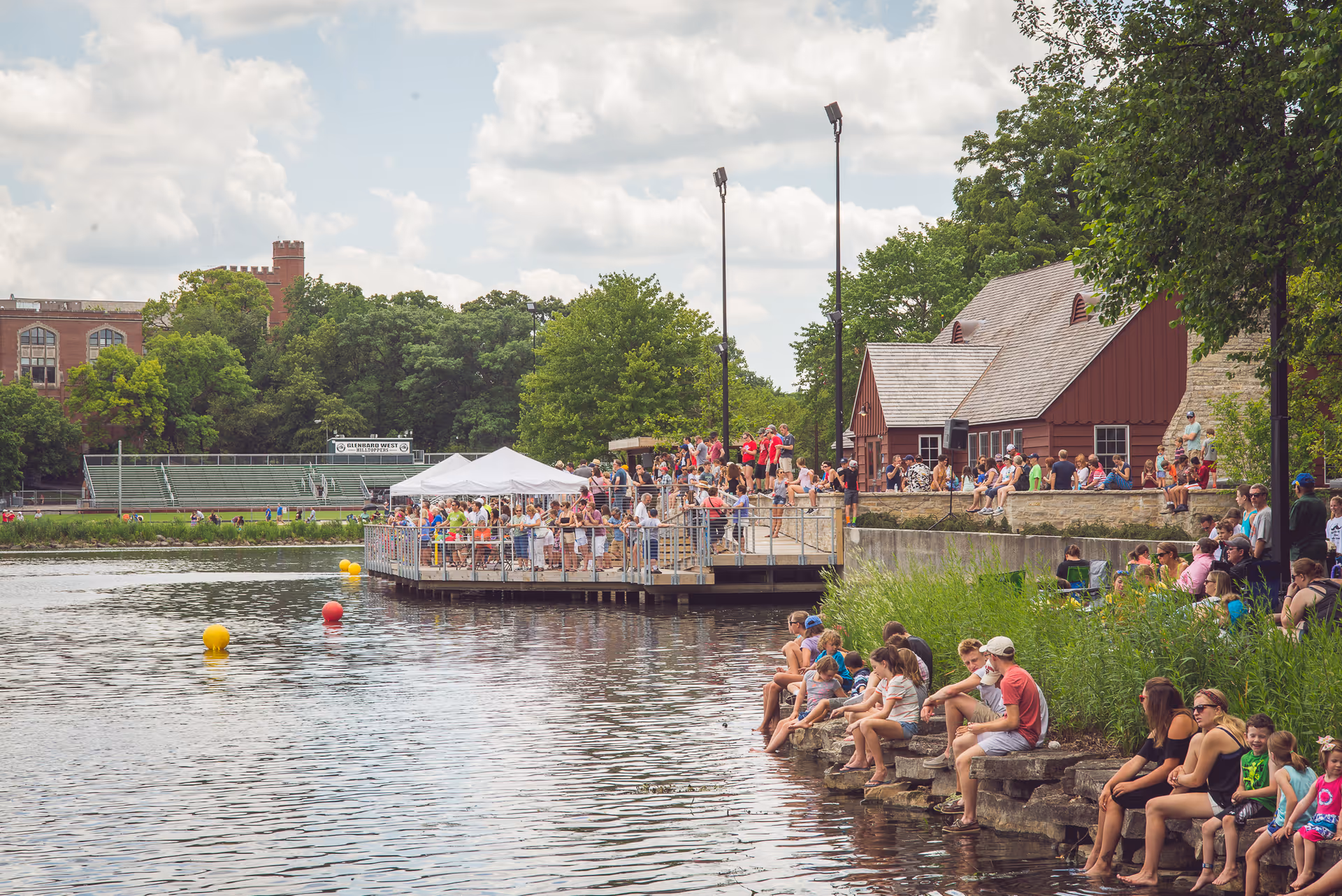 Crowd gathers on a lakeside dock with tents near red-roofed buildings.