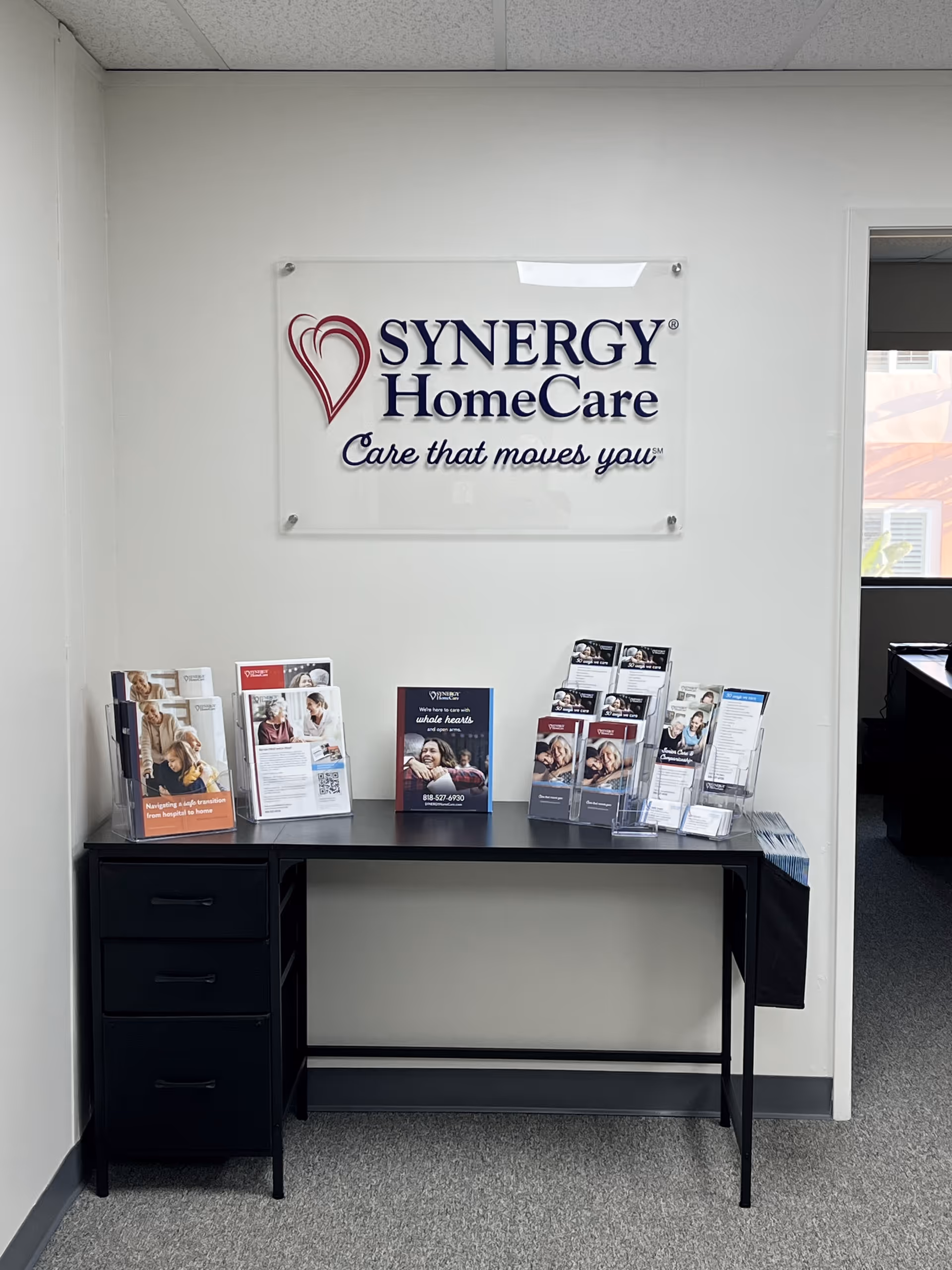 Interior reception area featuring a Synergy HomeCare sign and brochure display on a black desk.