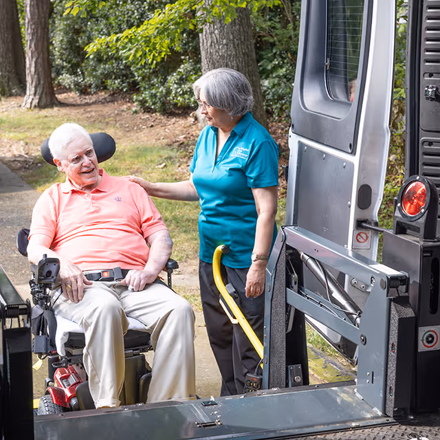 An elderly man in a wheelchair chats with a caregiver beside a mobility van outdoors.