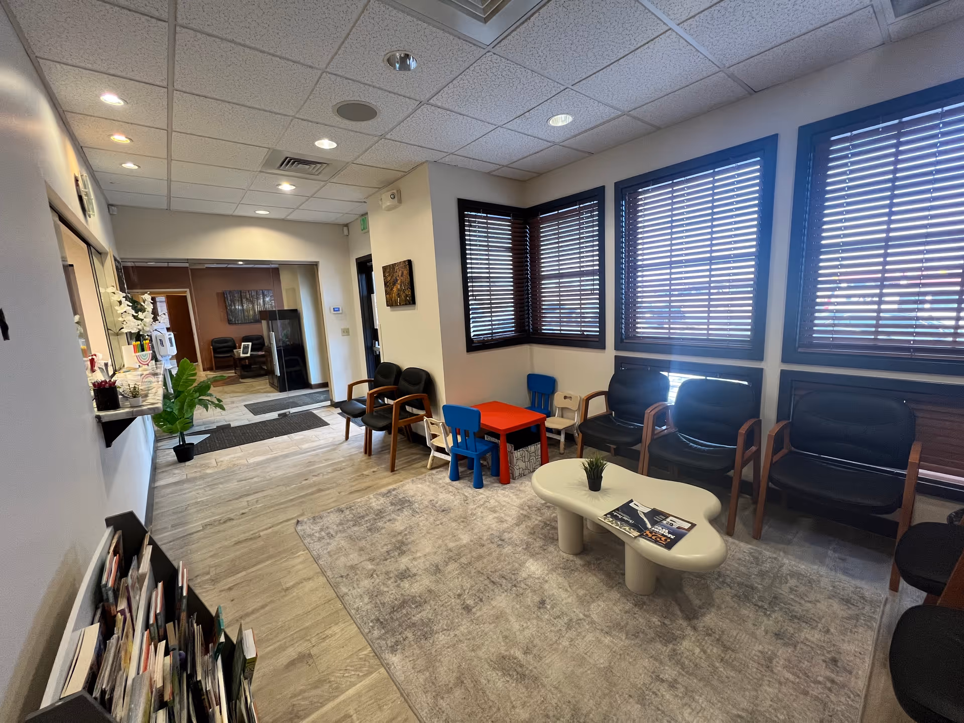Lobby-like waiting area with chairs, a child-sized table, magazines, plants, and large windows.