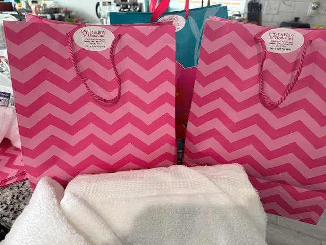Bright pink chevron gift bags labeled Synergy HomeCare sit on a counter with towels.