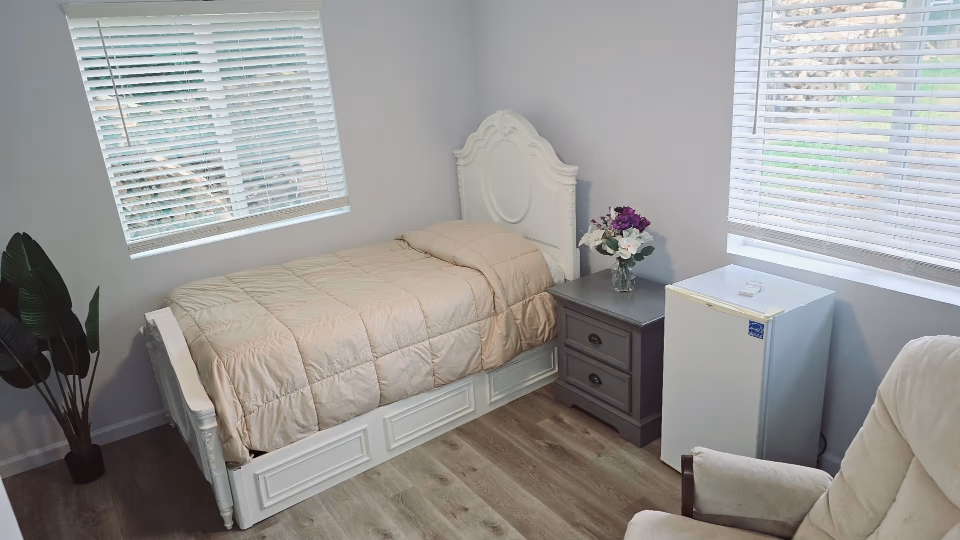 Cozy bedroom with a single ornate white bed, gray nightstand, mini fridge, and blinds.