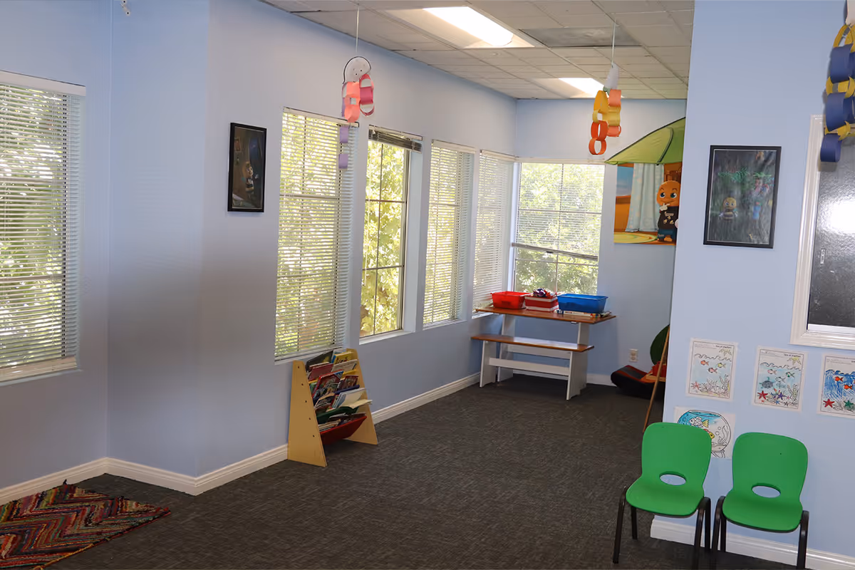 Light blue interior activity room with windows, colorful decorations, and child sized chairs around a table.