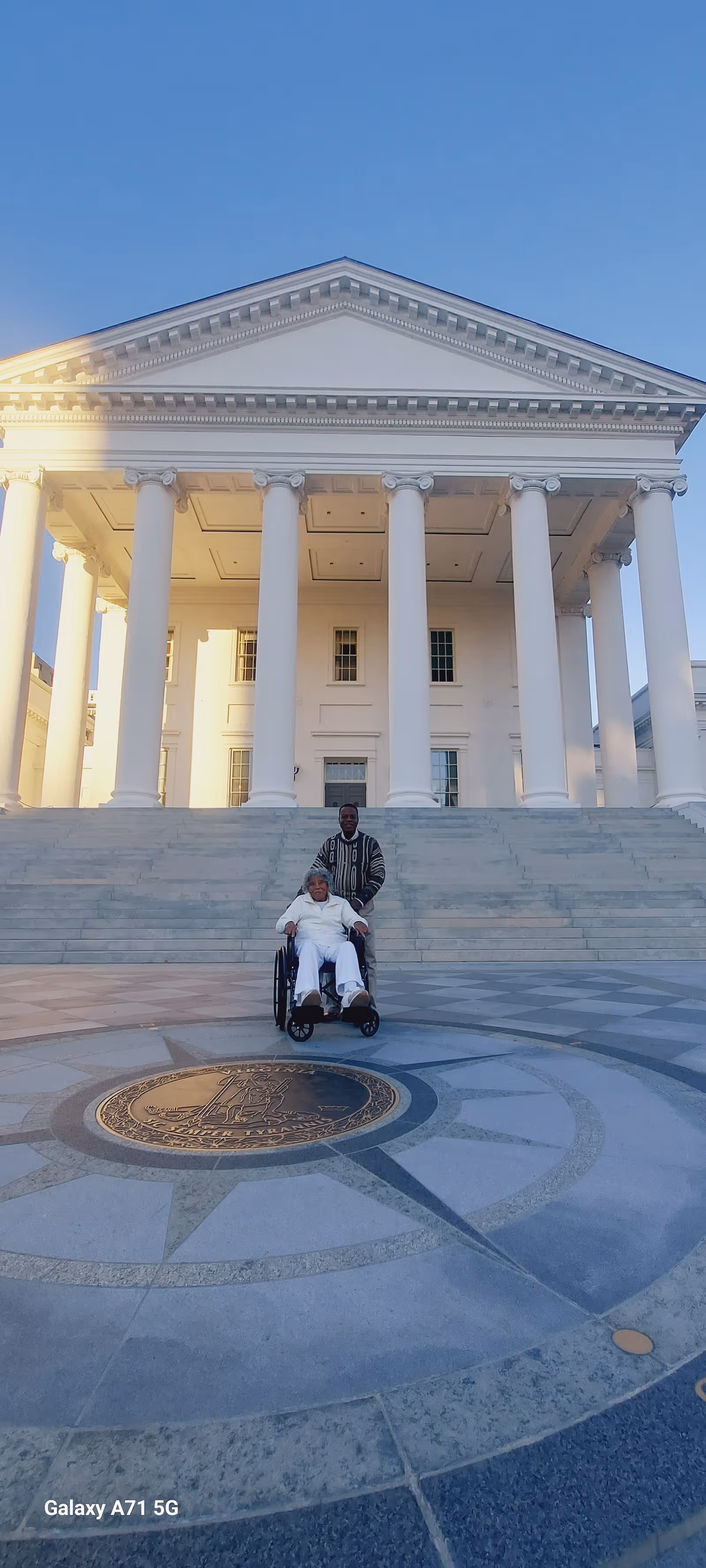 Two people, one in a wheelchair, pose on steps of a large white-columned building.
