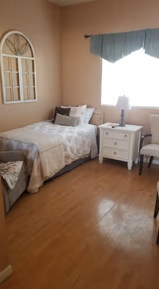 A cozy bedroom with a single bed, nightstand, lamp, and window in a senior living facility.