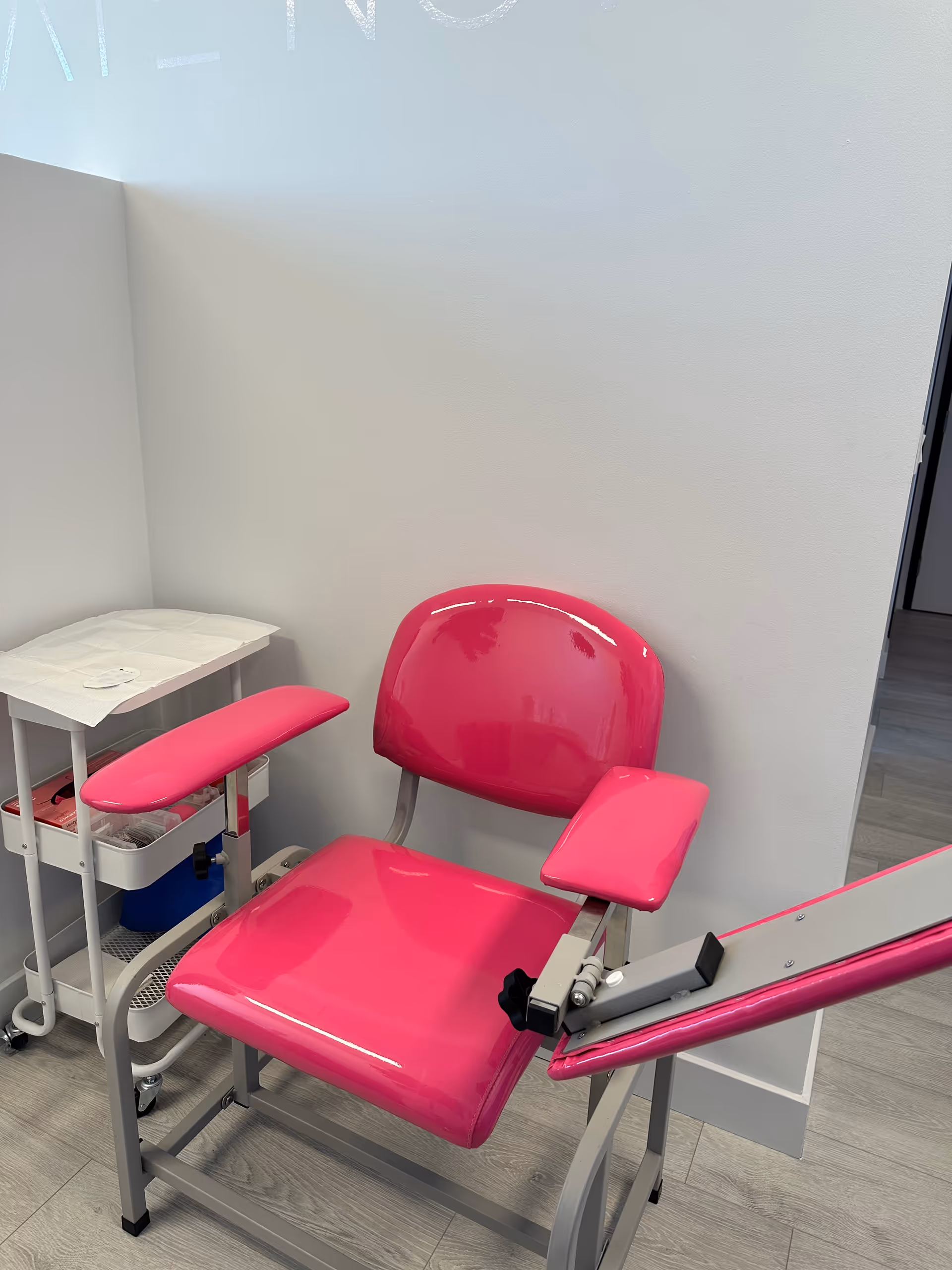 Bright pink examination chair with white cart in a small, white-walled clinic room.