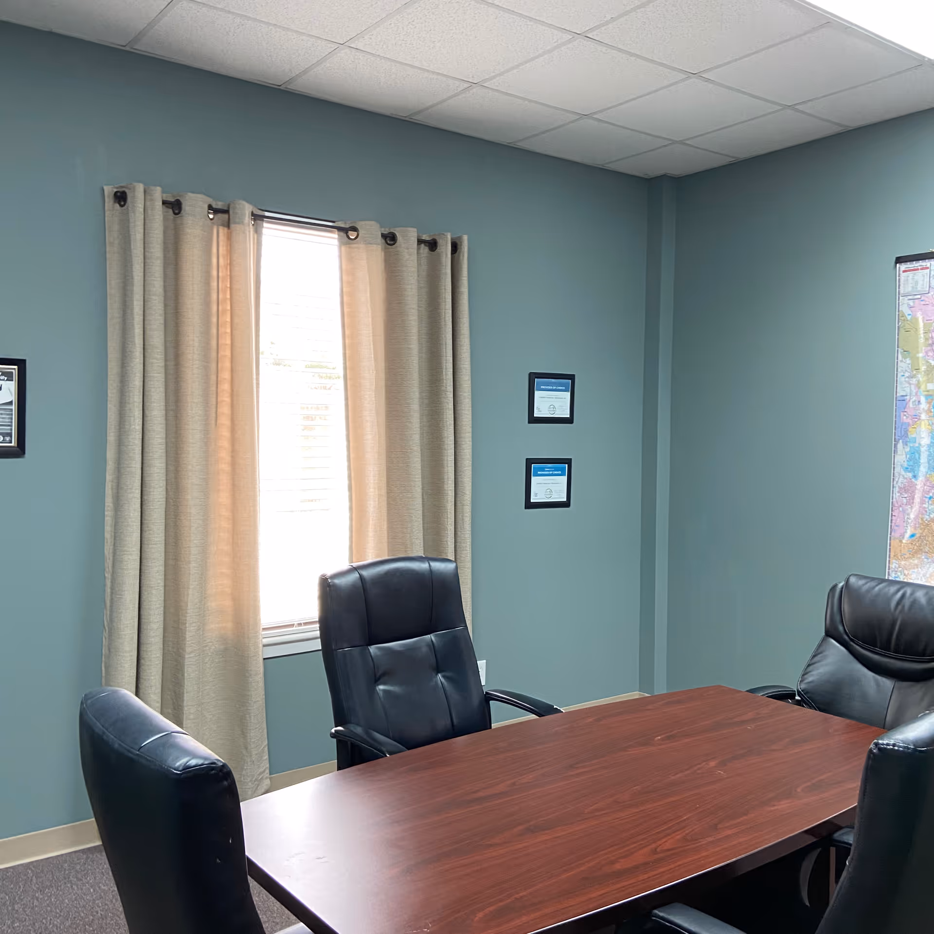 A conference room with a wooden table, black chairs, teal walls, window, and framed certificates.