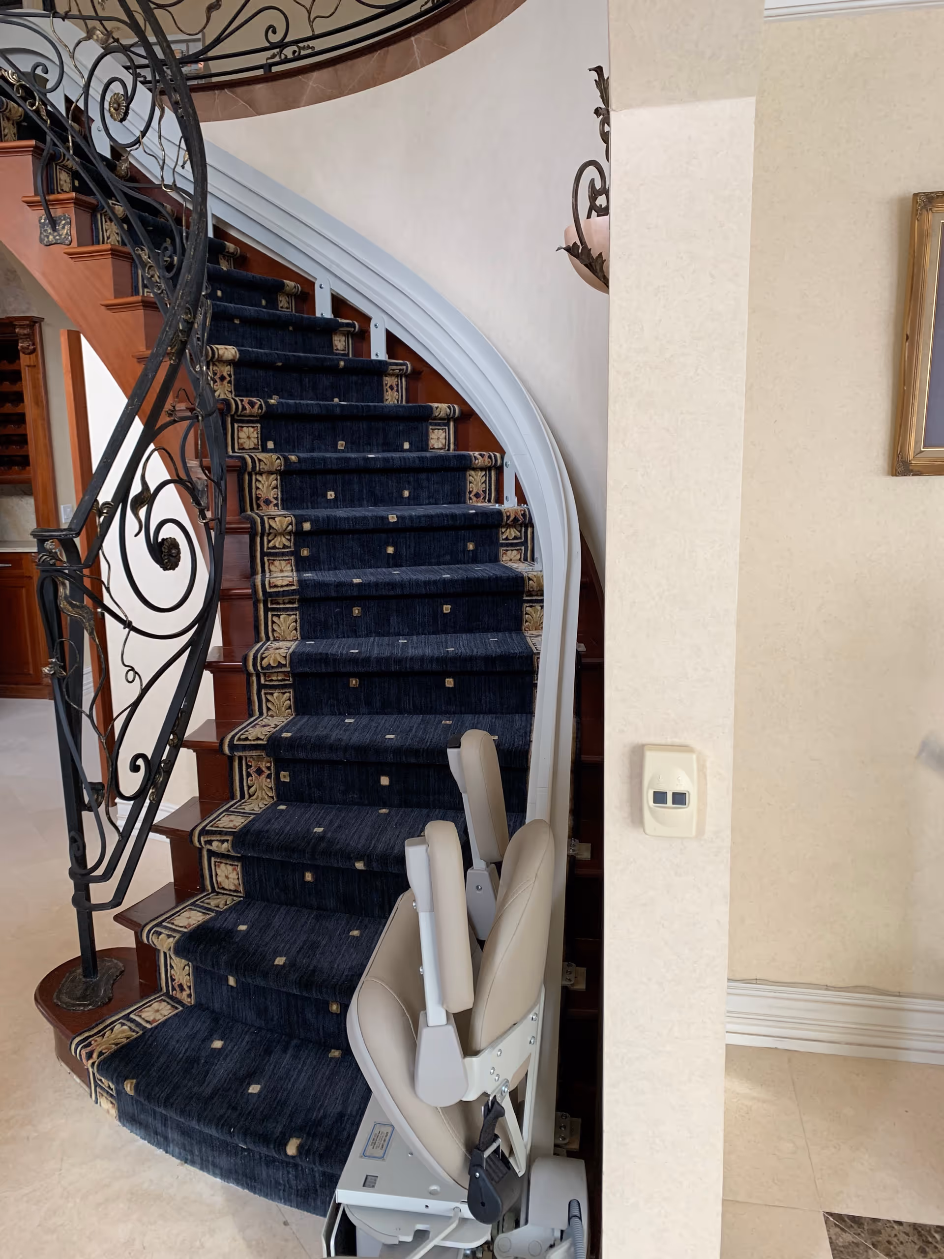 Curved staircase with ornate iron railing and beige stairlift chair inside a senior facility.