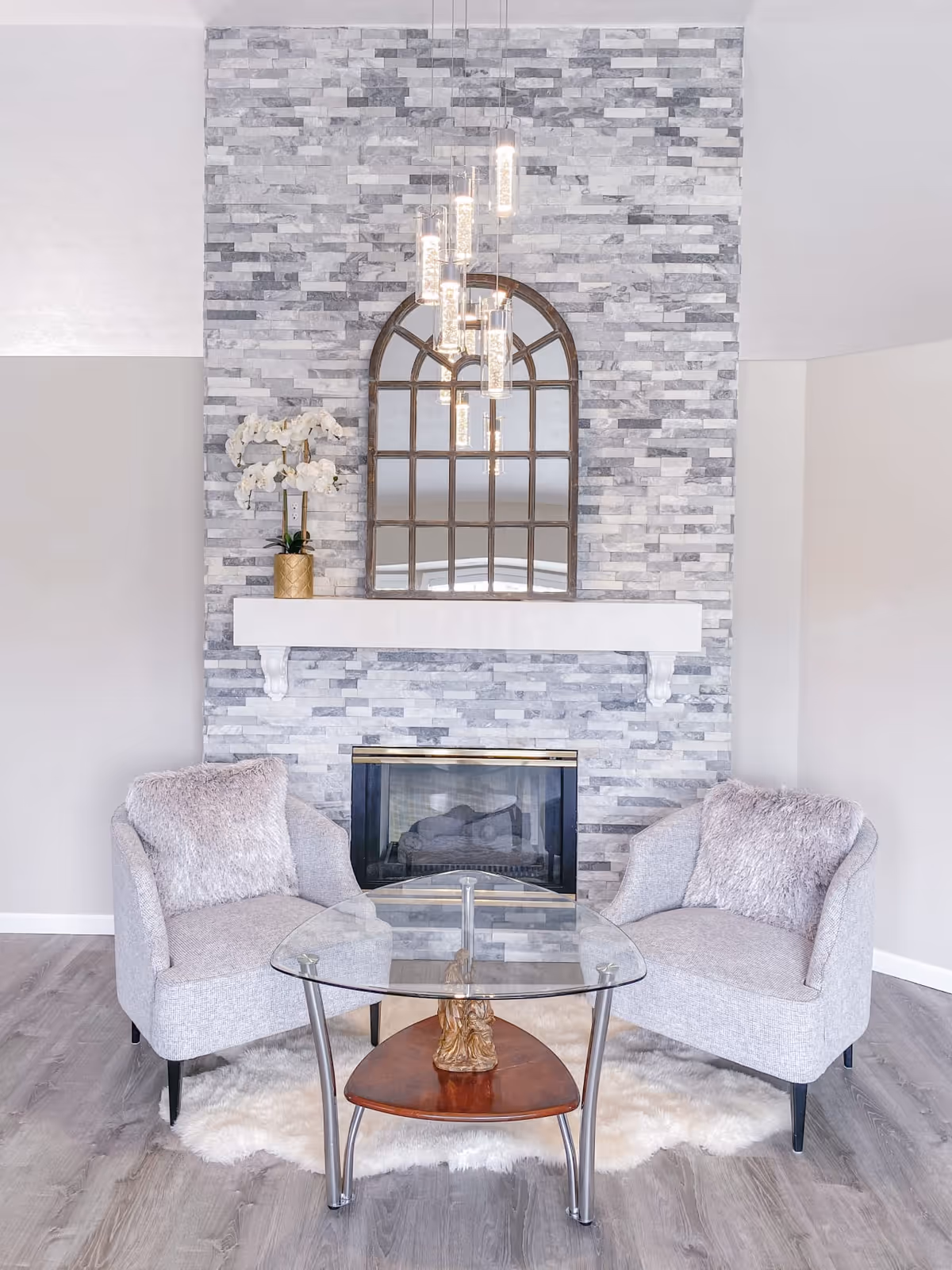 A cozy living room with a stone fireplace, arched mirror, two chairs, and glass table.