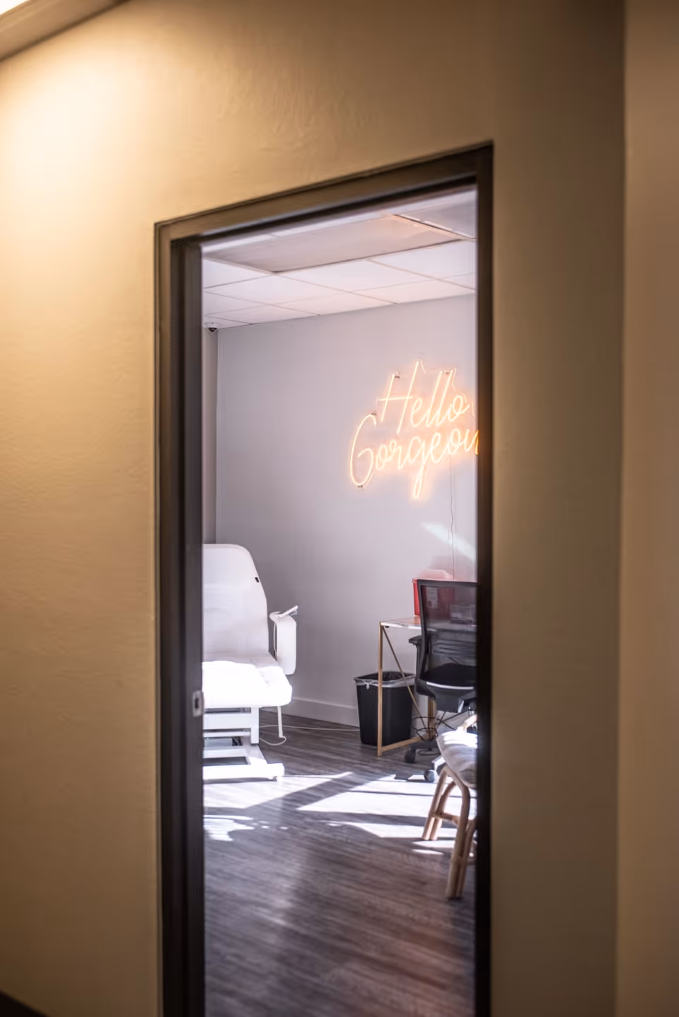 View through doorway of a treatment room featuring a neon Hello Gorgeous sign, chair, and desk.