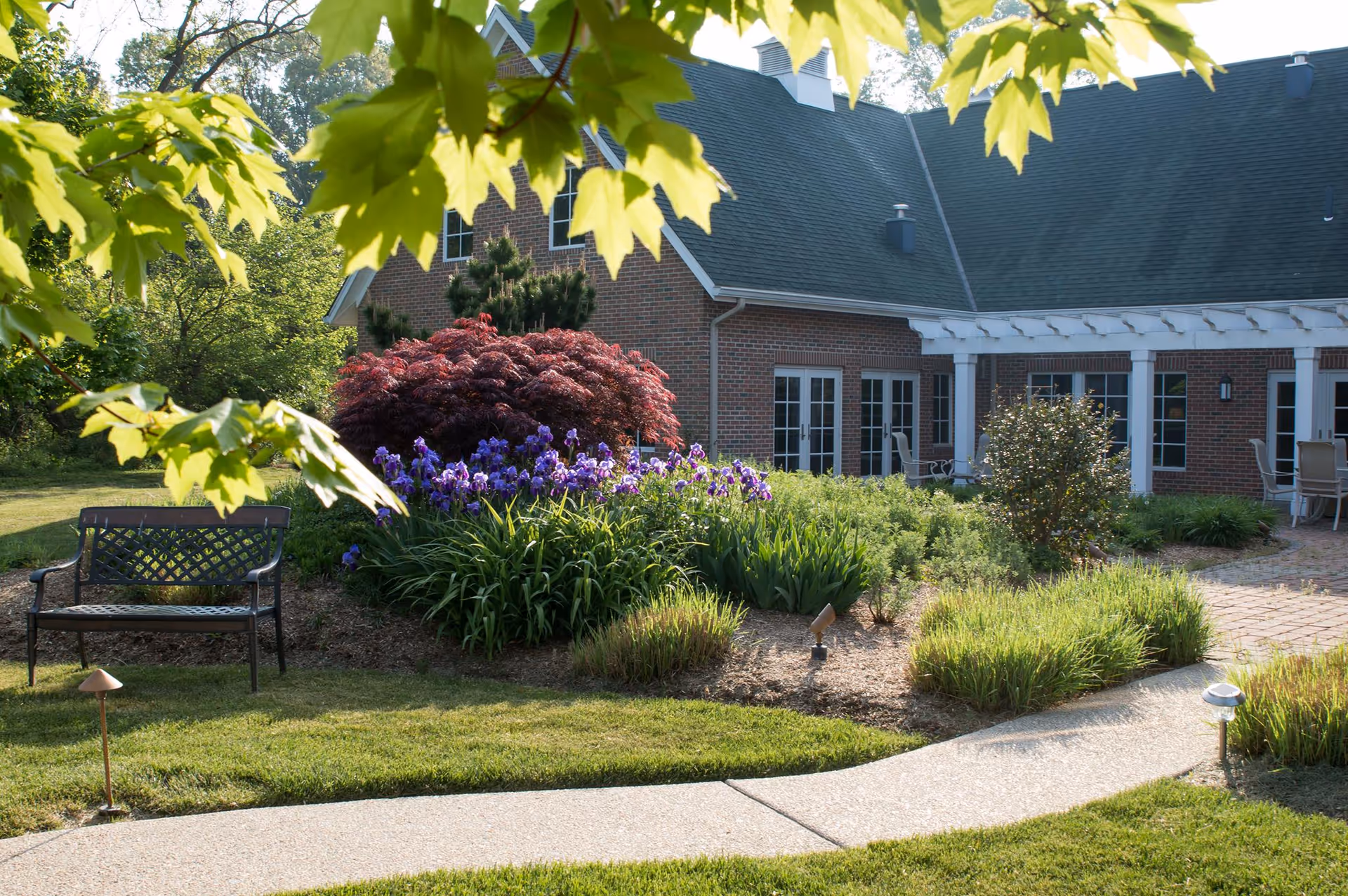 Brick senior living facility with manicured garden, bench, and patio near windows.
