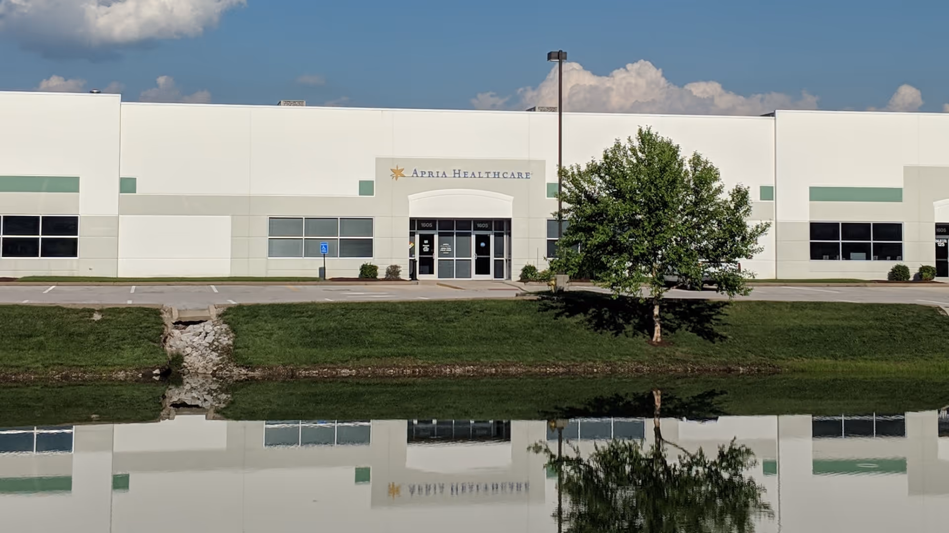 White healthcare building labeled Apria Healthcare with a tree, parking lot, and water reflection.