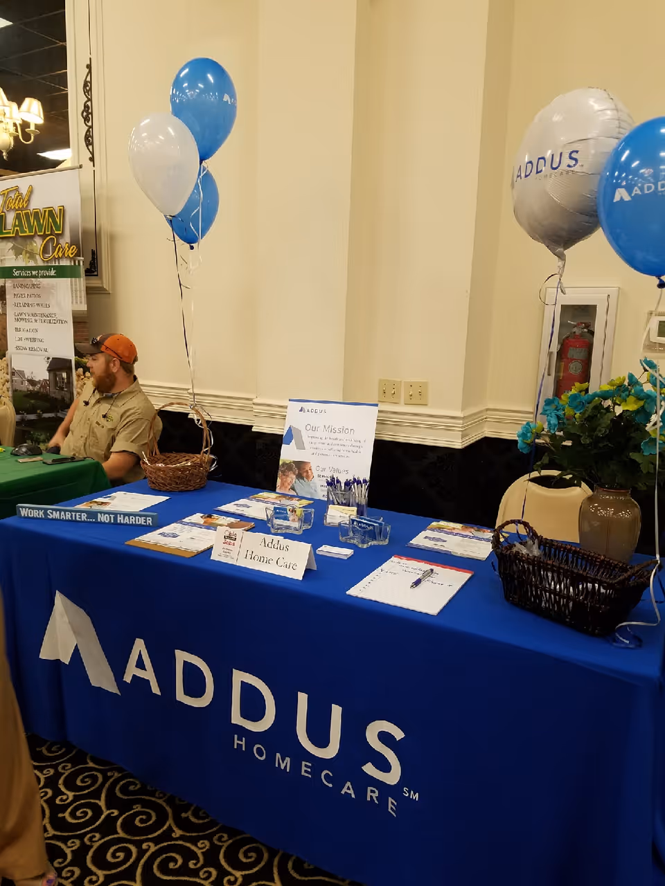 Indoor event booth for Addus Home Care with blue table, balloons, and a seated staff member.