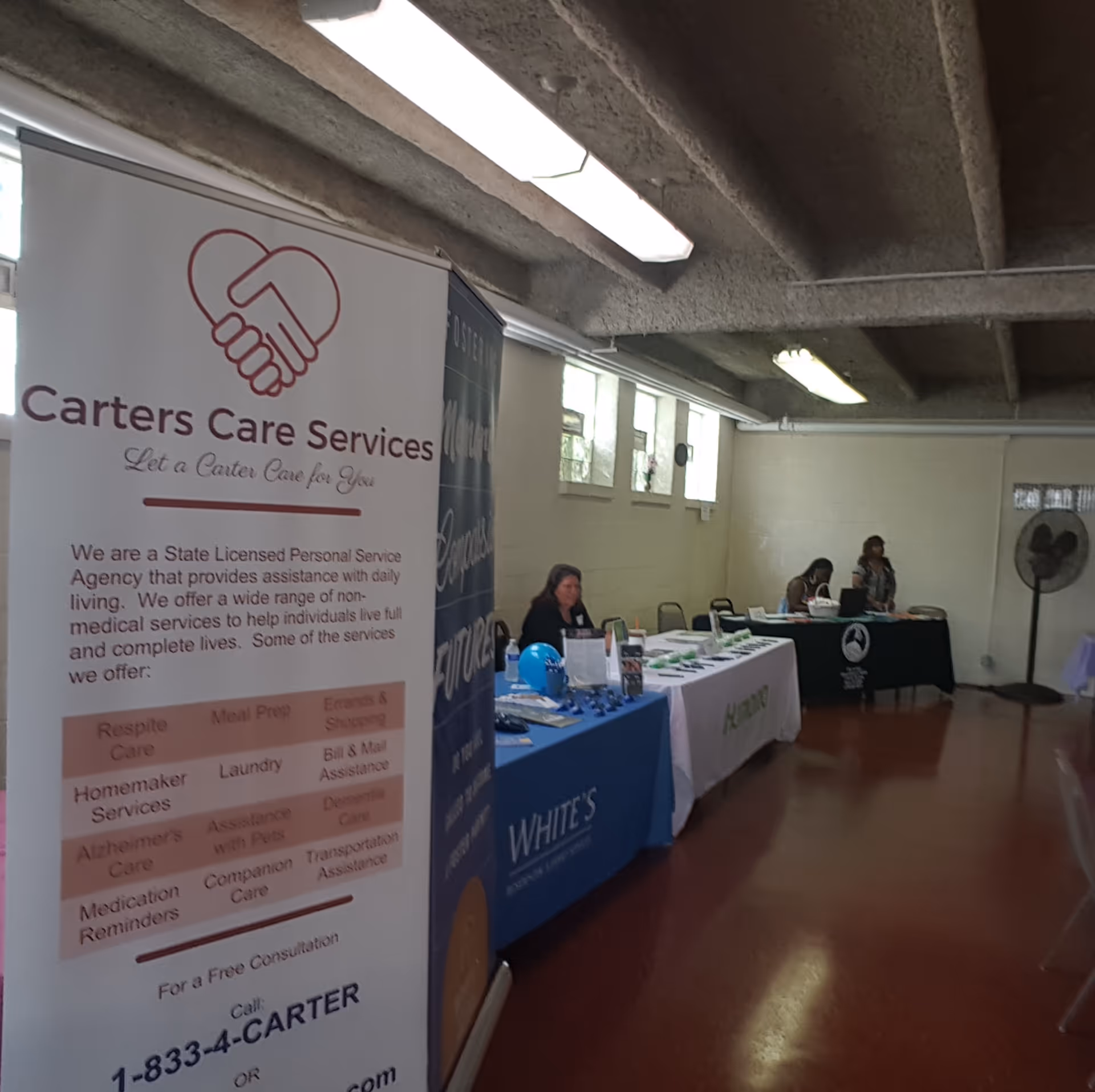 Indoor community fair setup with banners, tables, and volunteers in a care facility.