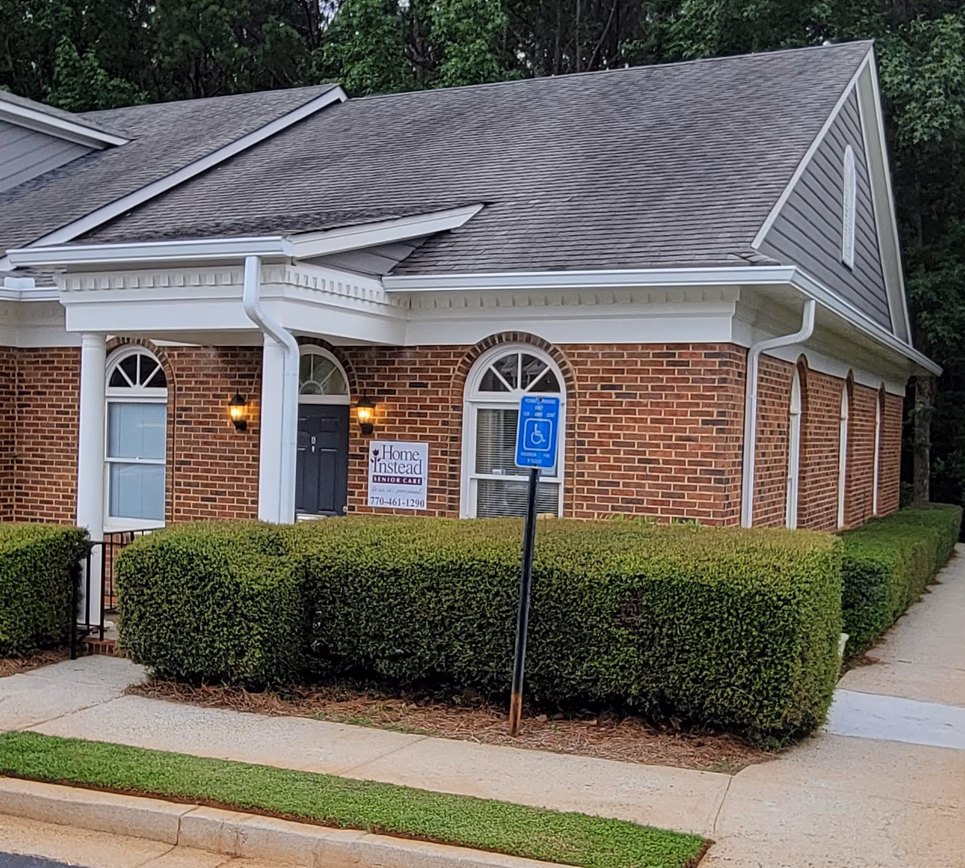 Brick senior living building with hedges, handicap sign, and Home Instead sign near entrance.