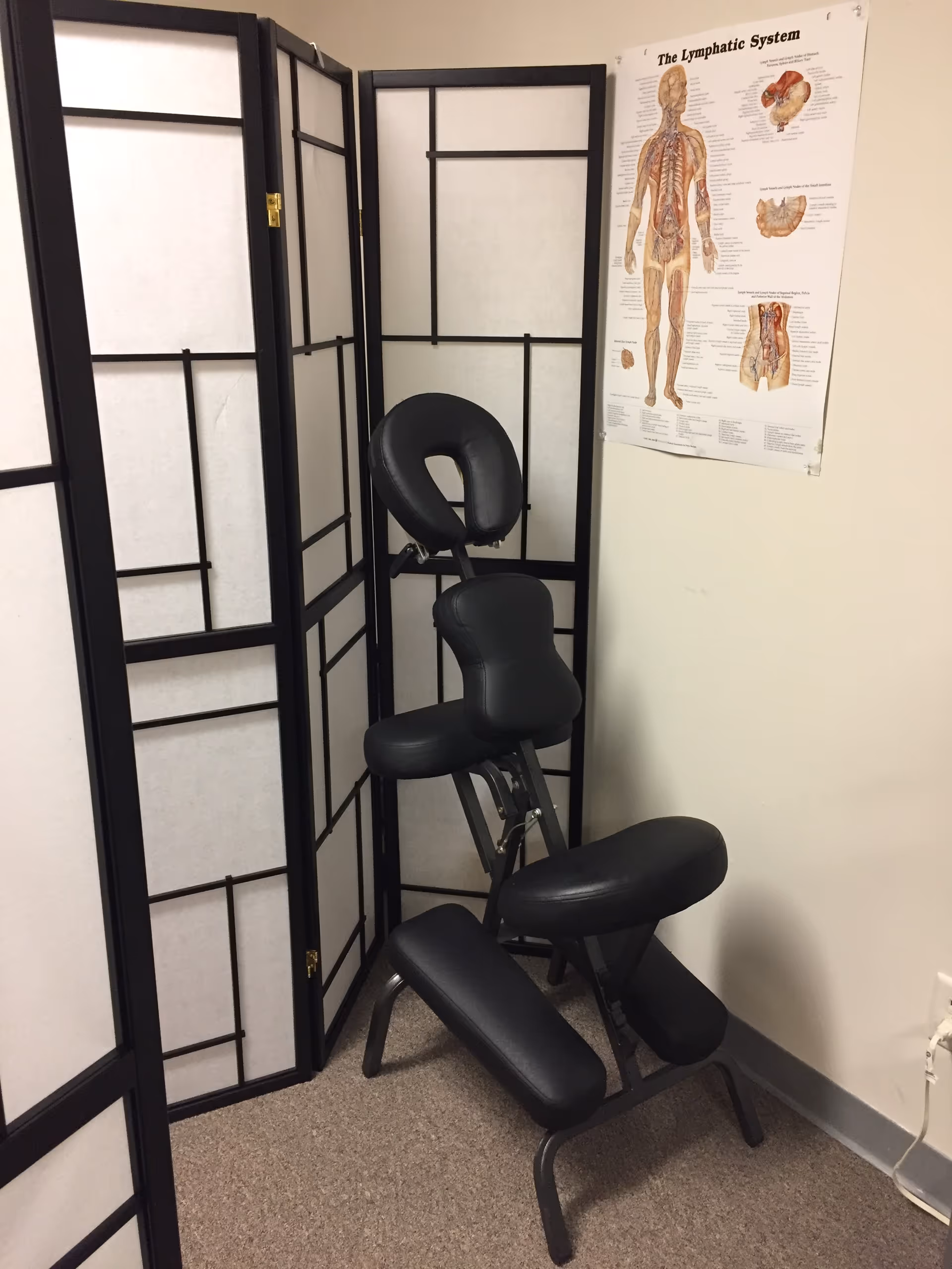 A black therapy chair beside folding privacy screens in a treatment room with a wall poster.