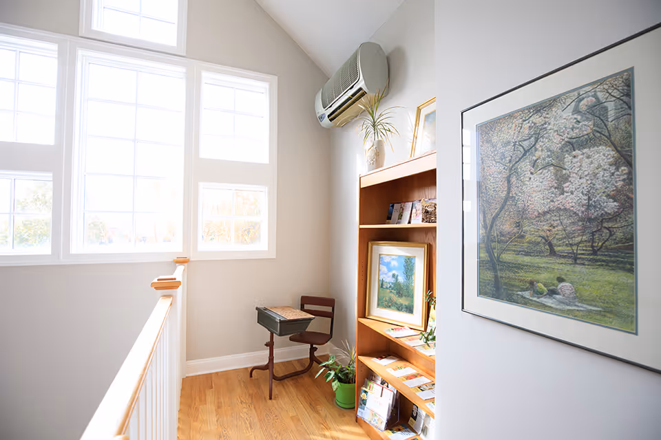 Bright upstairs hallway with large windows, a wooden bookshelf, plant, and framed art.