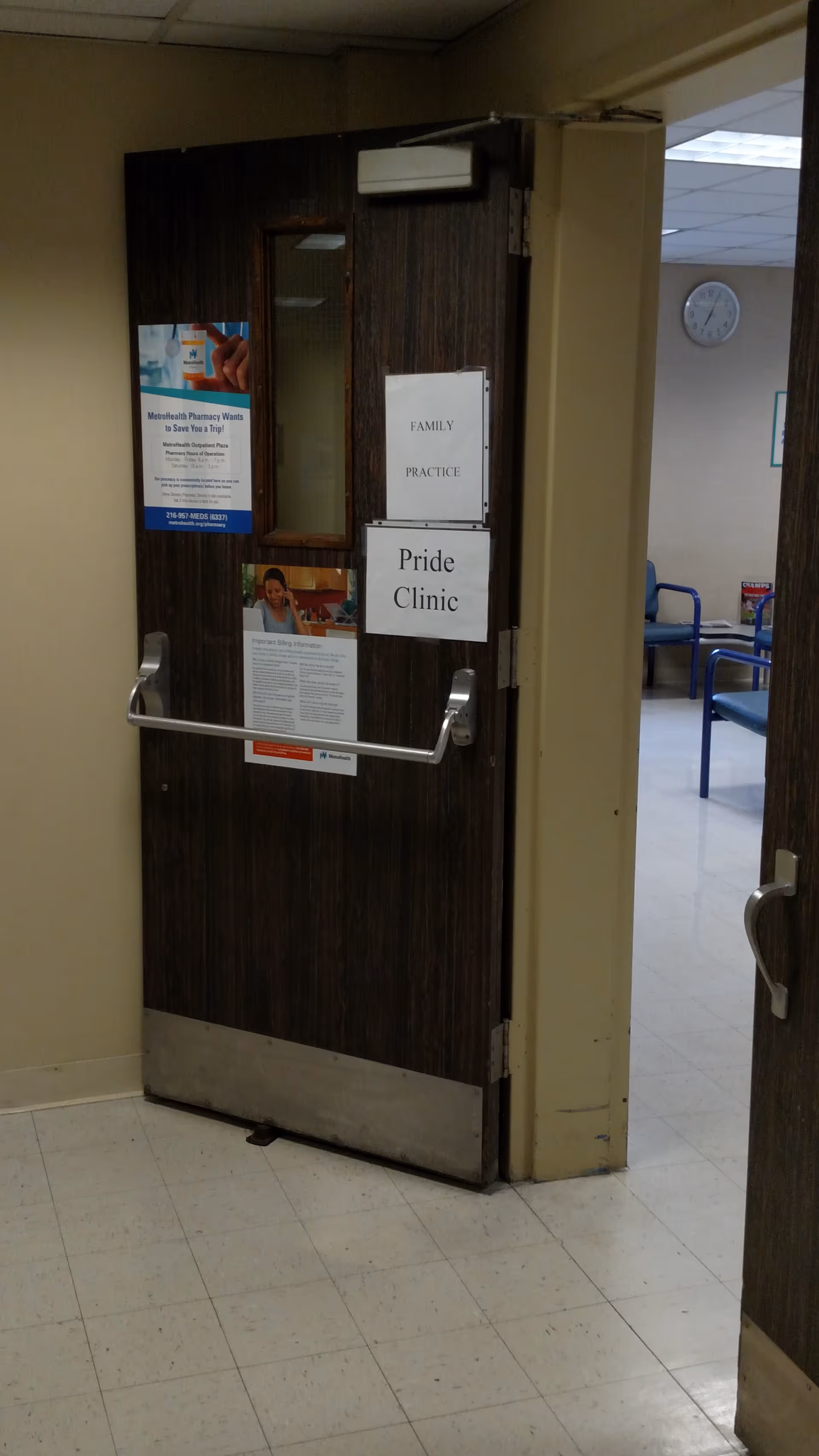 Wooden door in a beige hallway with Pride Clinic and Family Practice signs; chairs visible beyond.