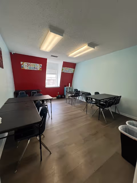 A classroom-style activity room with multiple tables, chairs, posters, and light blue walls with red accents.